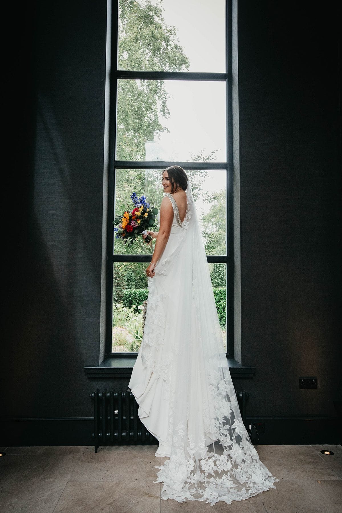 Bride standing at a tall window, holding her wedding flowers and glancing back